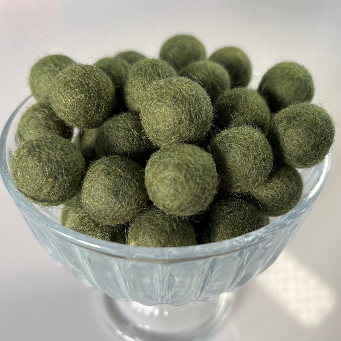 Olive green felt balls in a glass bowl on a white background