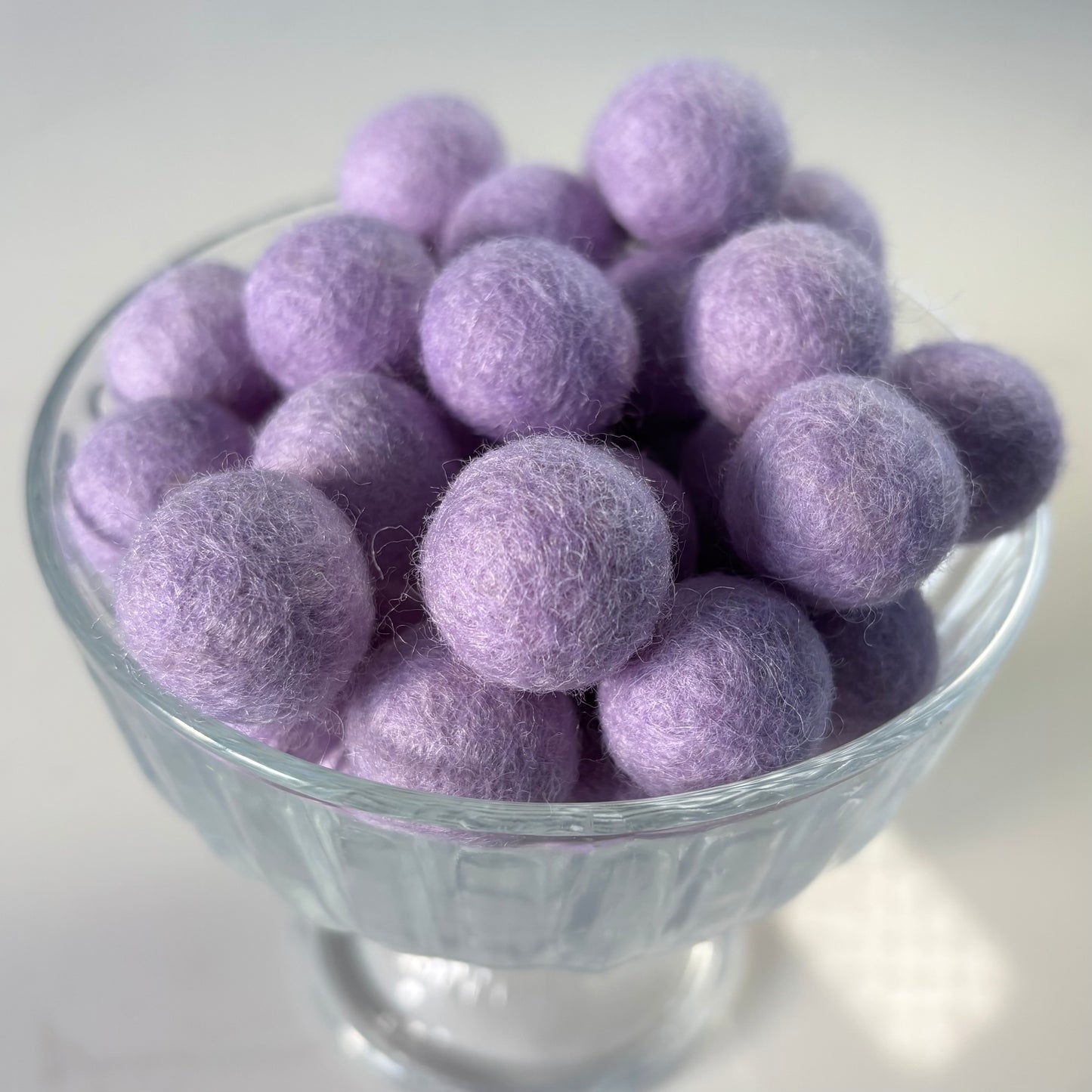 Glass bowl filled with light purple felt balls on a white background