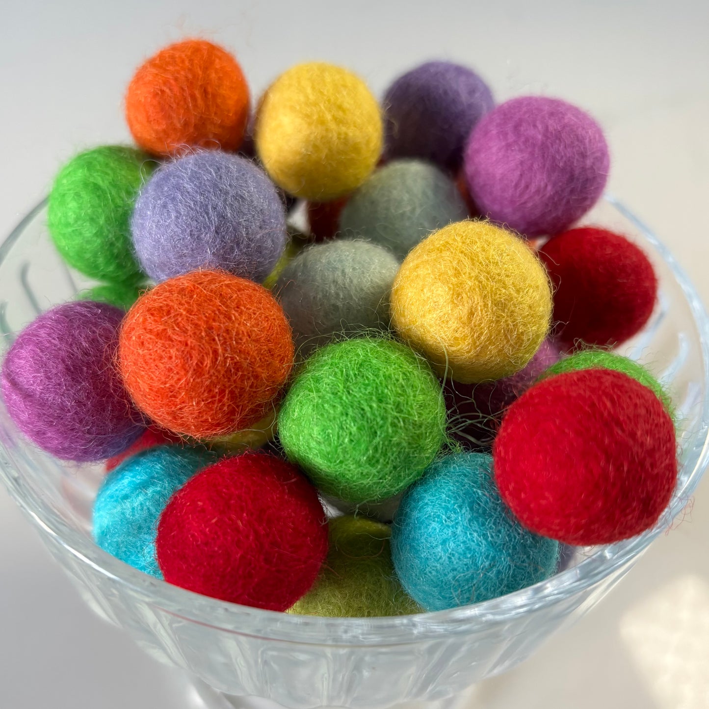 Colorful felt balls in a clear glass bowl on a white background