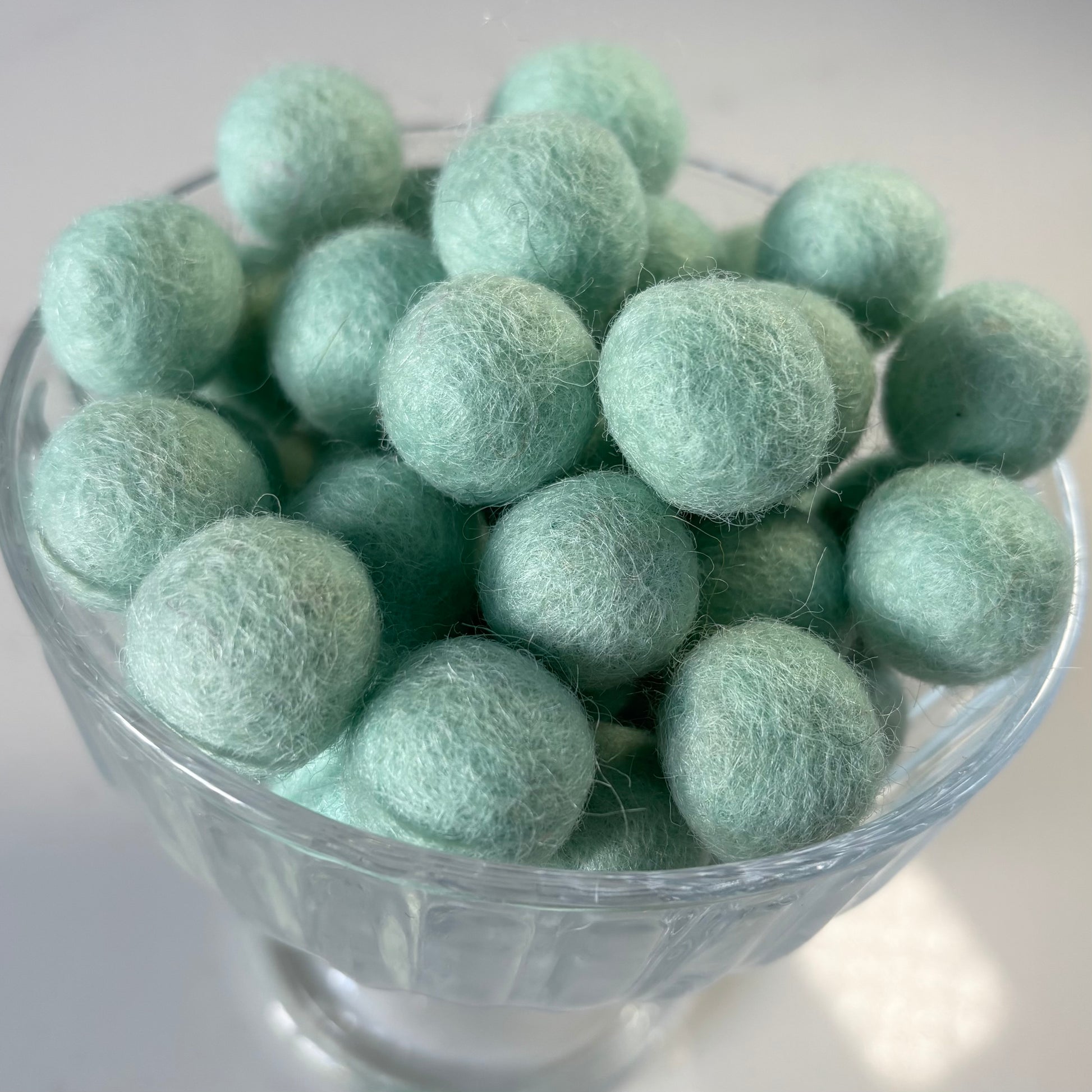 Glass bowl filled with light aqua green felt balls on a white background