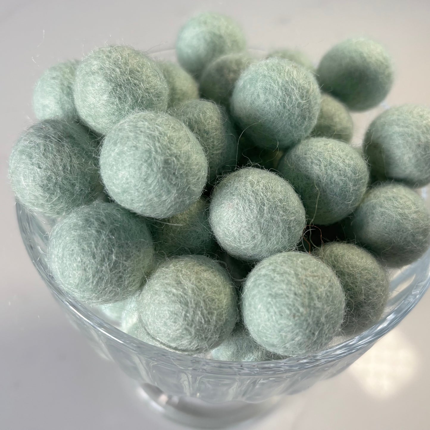 Glass bowl filled with light peppermint green felt balls on a white background