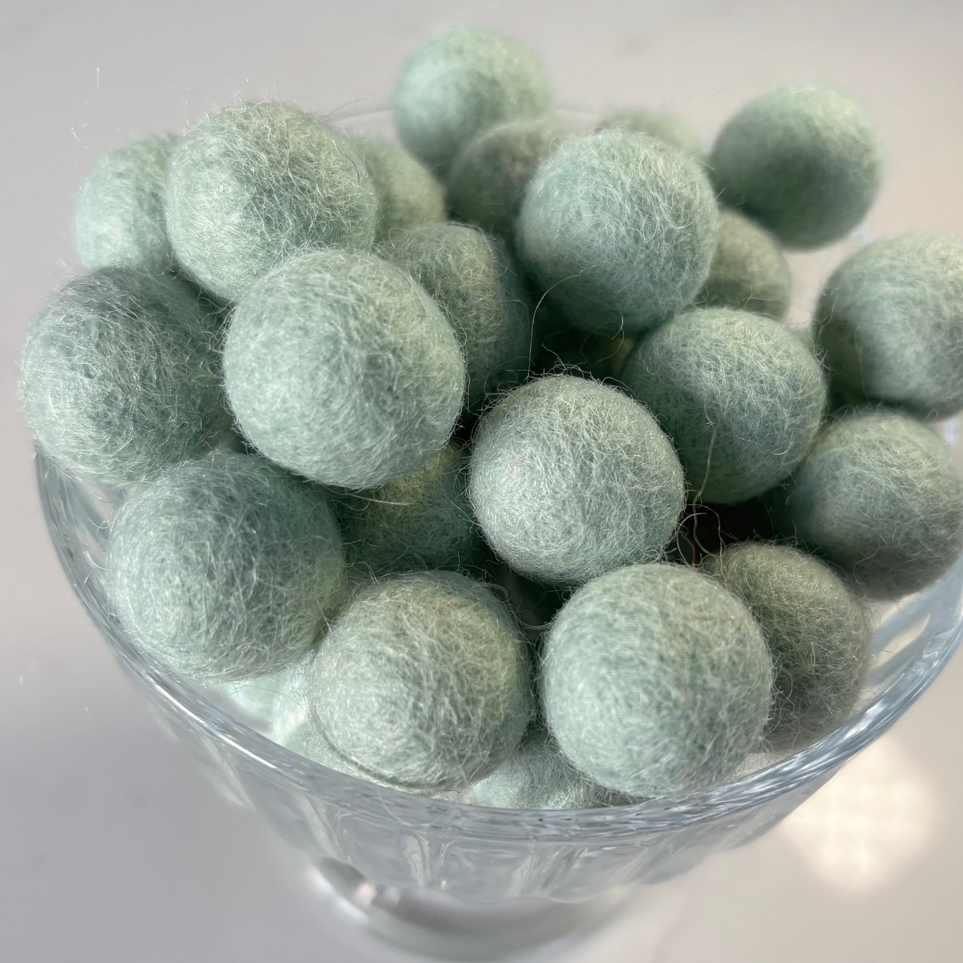 Glass bowl filled with light peppermint green felt balls on a white background