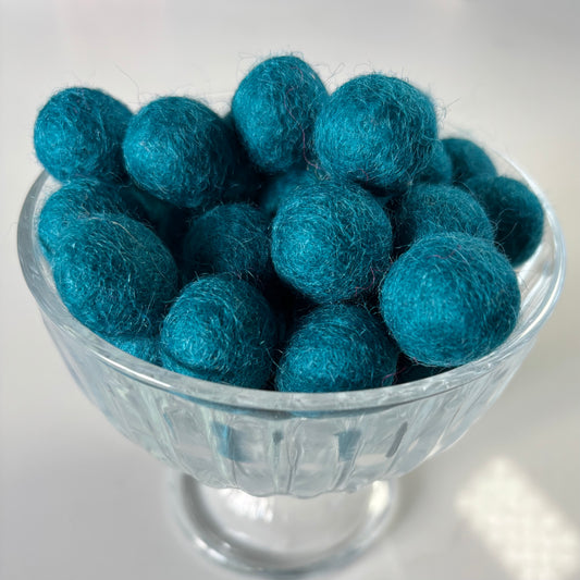 Teal felt balls in a glass bowl on a white background